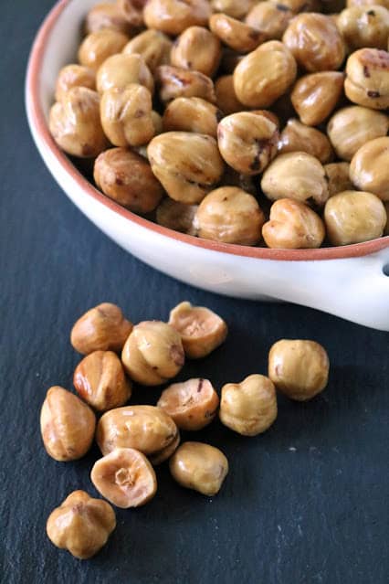 A white bowl filled with roasted hazelnuts sits on a dark surface, with several hazelnuts scattered outside the bowl in the foreground.