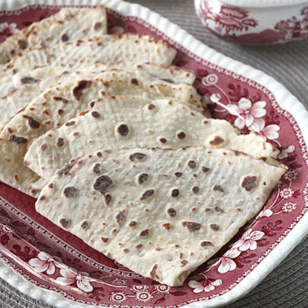 Several pieces of flatbread with brown spots are arranged on a decorative red and white floral-patterned plate, sitting on a woven placemat. A butter dish and spreader are visible in the background.