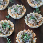 Mini phyllo pastry cups filled with a creamy mushroom mixture, garnished with fresh herbs, are arranged on a dark wooden surface.