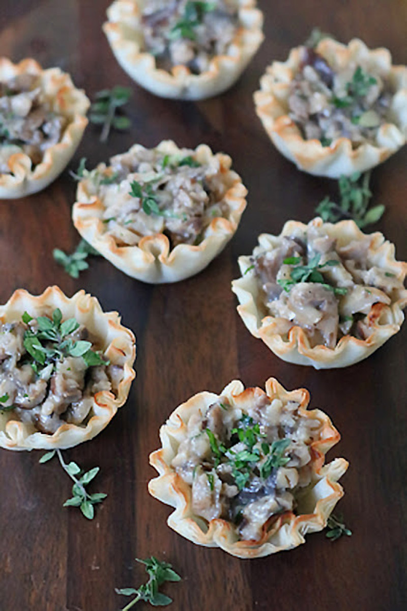 Mini phyllo pastry cups filled with a creamy mushroom mixture, garnished with fresh herbs, are arranged on a dark wooden surface.
