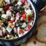 A bowl of chopped antipasto salad with olives, cheese, salami, and vegetables, surrounded by slices of toasted bread on a dark surface.