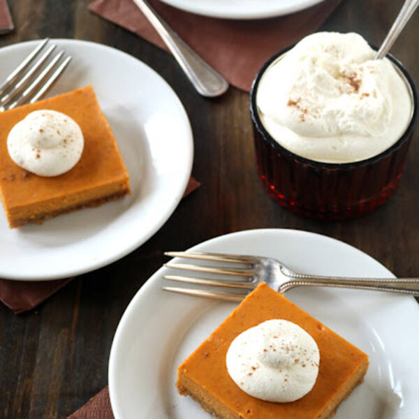 Three white plates each have a pumpkin pie bar topped with a dollop of whipped cream beside forks, and a small glass bowl is filled with whipped cream and a spoon on a dark wooden table.