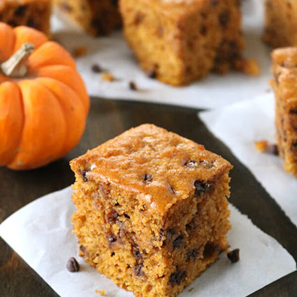 A square piece of pumpkin chocolate chip cake sits on parchment paper, with more pieces and a small pumpkin in the background on a wooden surface.