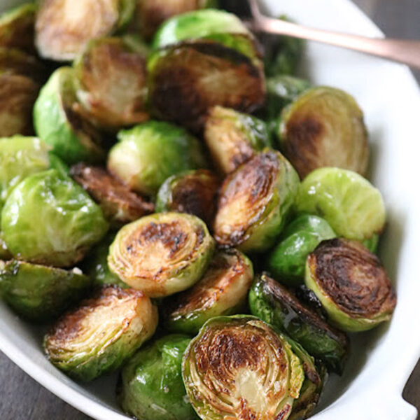 A white dish filled with roasted Brussels sprouts, some browned and crispy, with a serving spoon partially visible in the background.