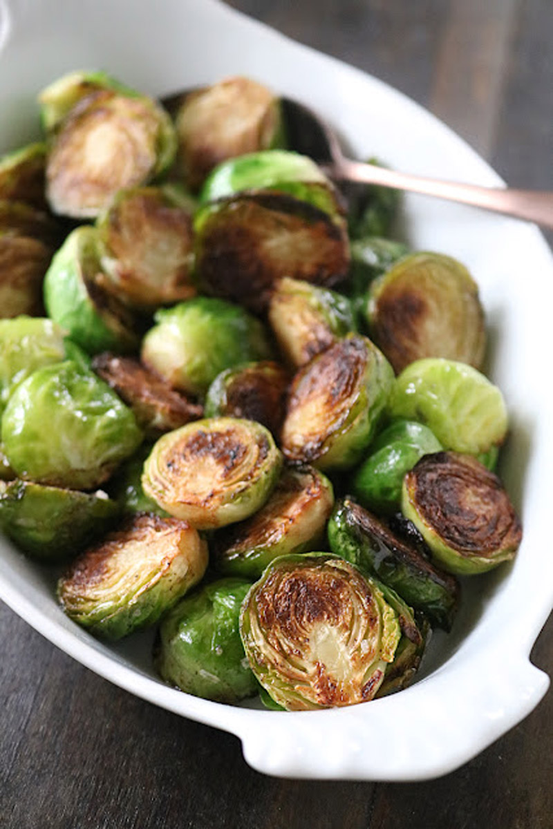A white dish filled with roasted Brussels sprouts, some browned and crispy, with a serving spoon partially visible in the background.