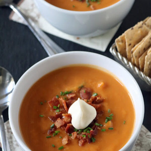 Two bowls of creamy orange soup topped with bacon bits, chopped chives, and a dollop of sour cream. Spoons, crackers, and napkins are placed nearby on the table.