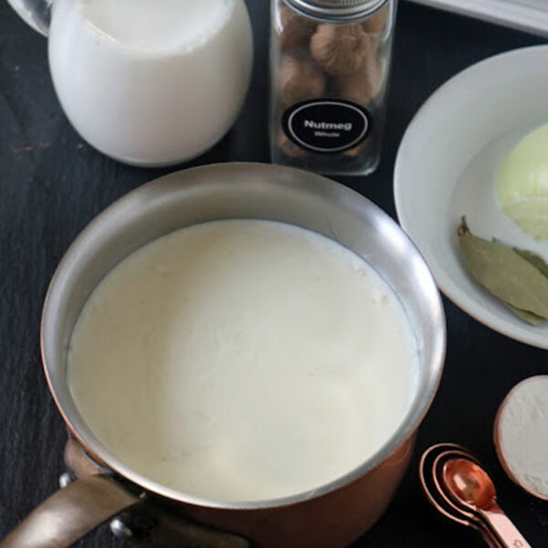 A saucepan filled with milk next to a small jug of milk, a jar of nutmeg, a white plate with onion and bay leaves, and copper measuring spoons on a dark surface.