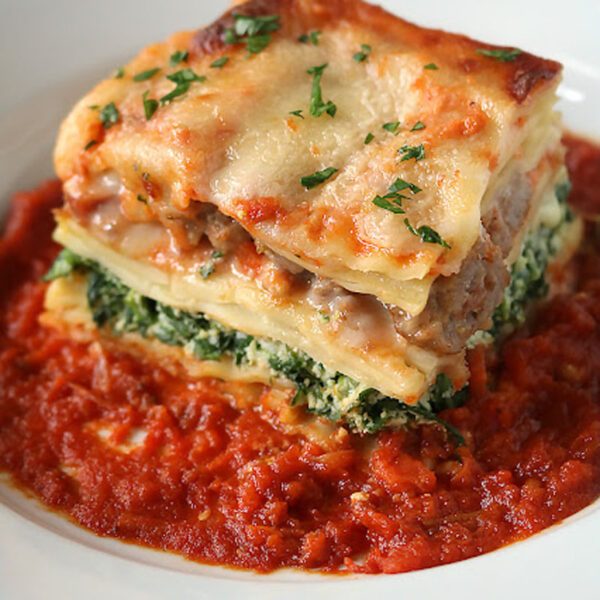 A slice of lasagna with layers of pasta, spinach, cheese, and meat sits on a plate, topped with melted cheese and herbs, and surrounded by a rich tomato sauce.