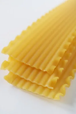 Close-up of several uncooked lasagna noodles arranged in a stack, showing their wavy, ruffled edges and ridged surface against a white background.