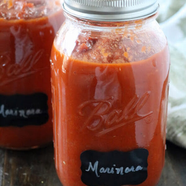 Two mason jars filled with red marinara sauce sit on a wooden surface. Each jar is sealed with a metal lid and labeled Marinara on a black label. A light cloth is in the background.