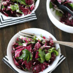 A fresh salad with radicchio and parsley leaves is served in white bowls on a dark wooden table, with forks placed on black-and-white striped napkins.