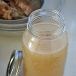 A glass mason jar filled with light brown homemade broth sits on a countertop with its lid off. In the background, a metal bowl holds cooked meat and bones.