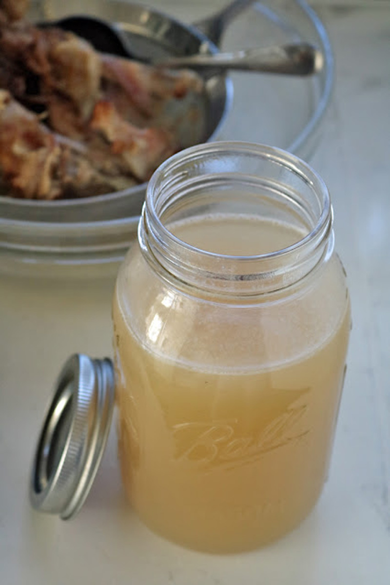 A glass mason jar filled with light brown homemade broth sits on a countertop with its lid off. In the background, a metal bowl holds cooked meat and bones.