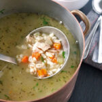 A pot of chicken soup with barley, carrots, celery, and herbs, with a metal ladle lifting a portion. In the background, a bowl of soup and two spoons rest on a folded napkin.