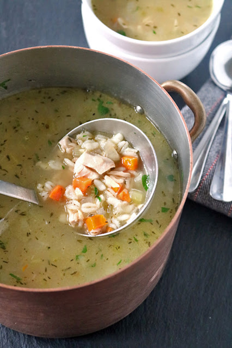 A pot of chicken soup with barley, carrots, celery, and herbs, with a metal ladle lifting a portion. In the background, a bowl of soup and two spoons rest on a folded napkin.