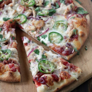 A baked pizza on a wooden board, topped with melted cheese, red onion, chopped cilantro, jalapeño slices, and pieces of chicken, with one slice being pulled away from the whole pizza.