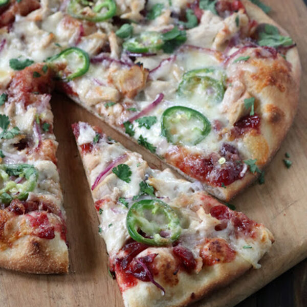 A baked pizza on a wooden board, topped with melted cheese, red onion, chopped cilantro, jalapeรฑo slices, and pieces of chicken, with one slice being pulled away from the whole pizza.