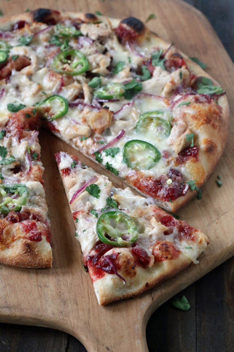 A baked pizza on a wooden board, topped with melted cheese, red onion, chopped cilantro, jalapeño slices, and pieces of chicken, with one slice being pulled away from the whole pizza.