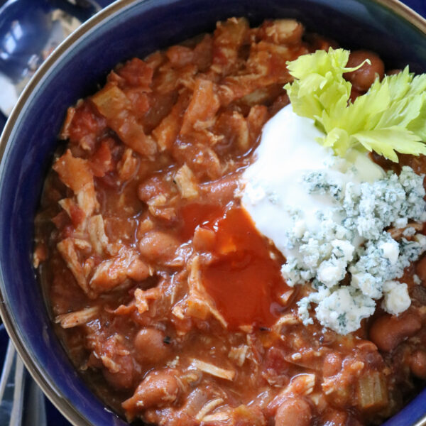 A bowl of chili topped with sour cream, blue cheese crumbles, celery, and hot sauce, with a spoon and crackers in the background.