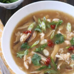 A bowl of hot and sour soup with chicken, rice, mushrooms, sliced green onions, cilantro, and drops of red chili sauce, served with a spoon and a side dish of fresh cilantro.
