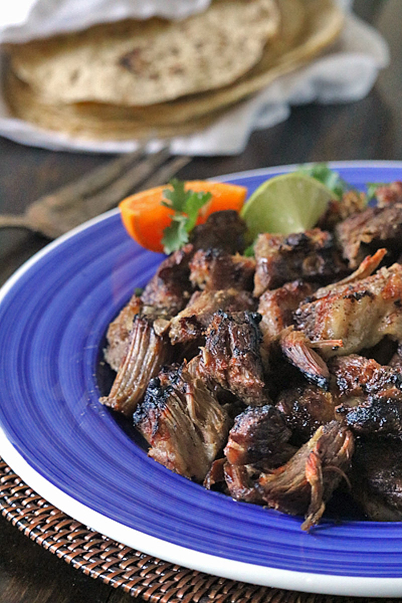 A blue plate filled with pieces of cooked, shredded beef garnished with cilantro, an orange wedge, and a lime wedge; a stack of tortillas is visible in the background.