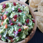 A wooden bowl of guacamole mixed with chopped red onion, cilantro, and pomegranate seeds, with tortilla chips in the background.
