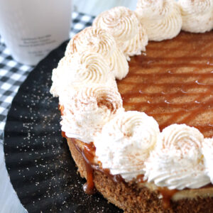 A close-up of a cheesecake topped with swirls of whipped cream and caramel drizzle on a black platter, with a bottle of RumChata in the background and a checkered cloth underneath.