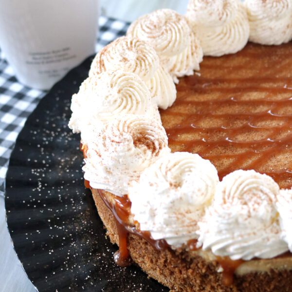 A close-up of a cheesecake topped with swirls of whipped cream and caramel drizzle on a black platter, with a bottle of RumChata in the background and a checkered cloth underneath.