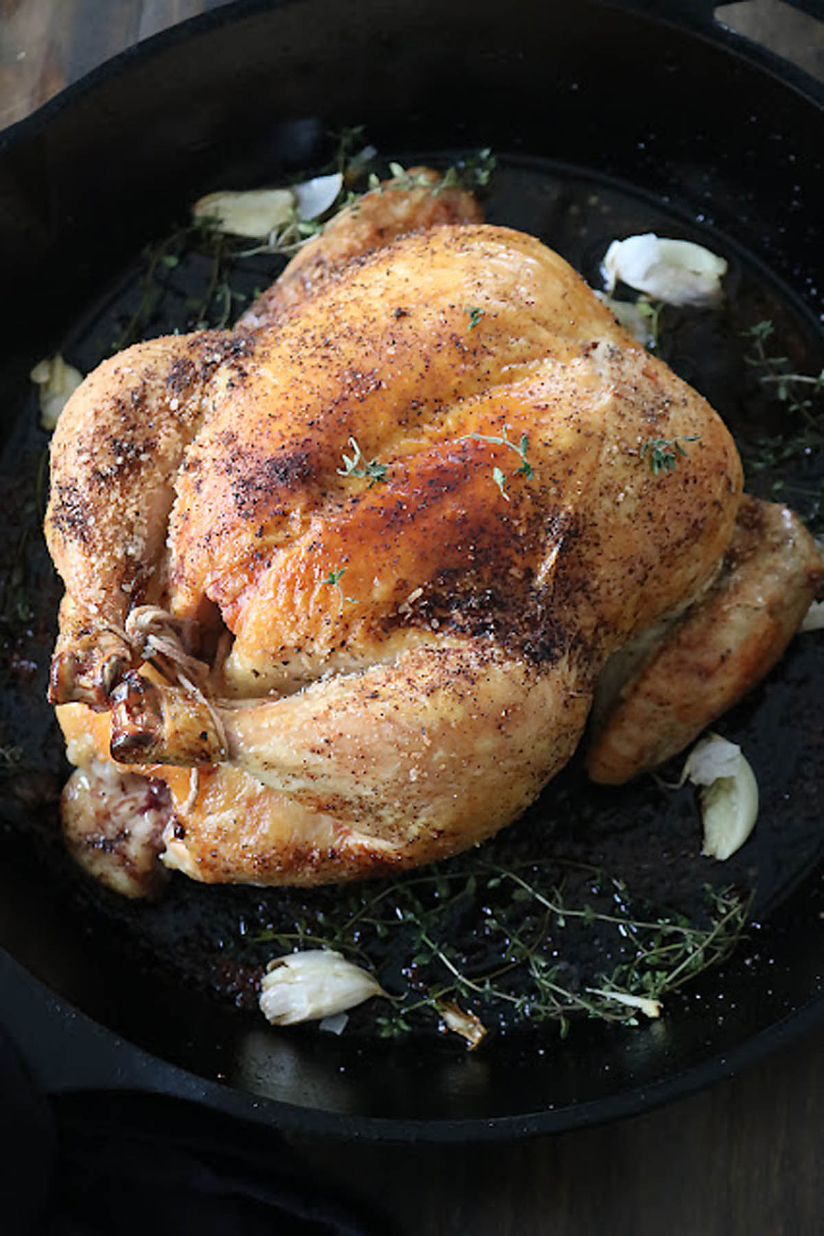 A whole roasted chicken in a black pan, garnished with fresh thyme sprigs and garlic cloves, with golden brown, crispy skin.