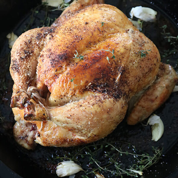 A whole roasted chicken seasoned with herbs sits in a black pan, surrounded by sprigs of fresh thyme and scattered garlic cloves.