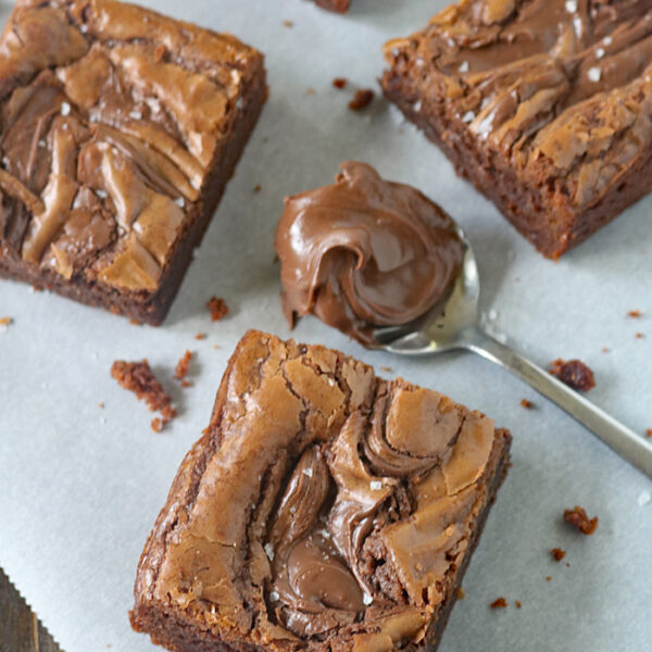 Three thick, fudgy brownies with a swirled chocolate topping sit on parchment paper. A spoon with a dollop of creamy chocolate spread is placed beside them. Brownie crumbs are scattered around.