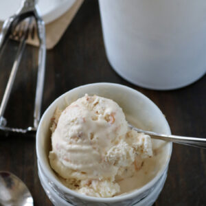 A bowl of creamy ice cream with a spoon rests on a dark wooden surface. In the background, there is an ice cream container and a metal scoop with traces of ice cream on a lid.
