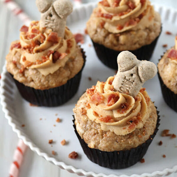 Four cupcakes topped with swirled frosting, crumbled bacon, and bone-shaped dog treats, displayed on a white plate. Red and white striped treats and bone-shaped biscuits are scattered in the background.
