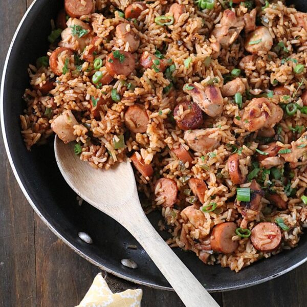 A skillet filled with rice, sliced sausage, chicken pieces, and green onions sits on a wooden table. A wooden spoon rests in the pan, and a yellow patterned napkin is nearby.
