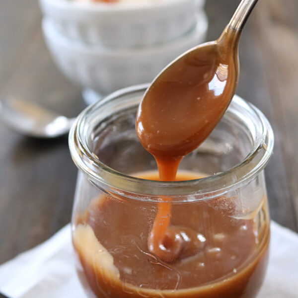 A spoon drizzles caramel sauce into a glass jar, with a bowl of ice cream topped with caramel in the blurred background.