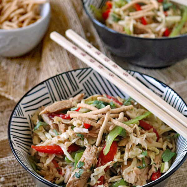 A bowl of Asian-inspired salad with sliced chicken, red bell peppers, cabbage, and crispy noodles, garnished with greens. A pair of chopsticks rests on top, with another bowl and a small dish of crispy noodles nearby.