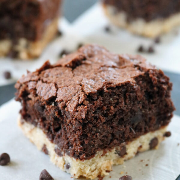 A close-up of a thick brownie square with a chocolate chip cookie crust, resting on parchment paper, with a few chocolate chips scattered around and more brownie squares in the background.