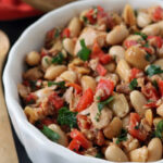 A white bowl filled with a salad made of white beans, chopped red peppers, parsley, and pieces of tuna. The ingredients are mixed together, with a wooden spoon and bread visible in the background.