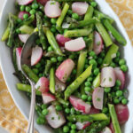 A white dish filled with a colorful vegetable medley of green peas, asparagus, and sliced radishes, garnished with herbs. A silver serving spoon rests in the dish, which sits on a yellow patterned napkin.