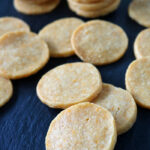 Round, golden-brown crackers are scattered and stacked in small groups on a dark, slate surface. The crackers appear crispy with a slightly textured surface.