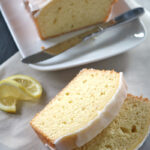 Two slices of glazed lemon pound cake rest on parchment paper beside a partially sliced loaf on a white plate. A knife and lemon wedges are placed nearby. The cake has a light, moist texture.