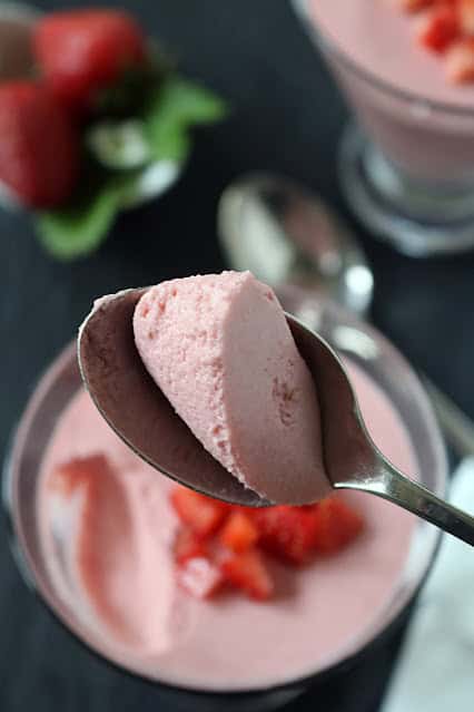A close-up of a spoon holding a scoop of light pink strawberry mousse above a glass bowl, with fresh strawberry pieces and another dessert cup blurred in the background.