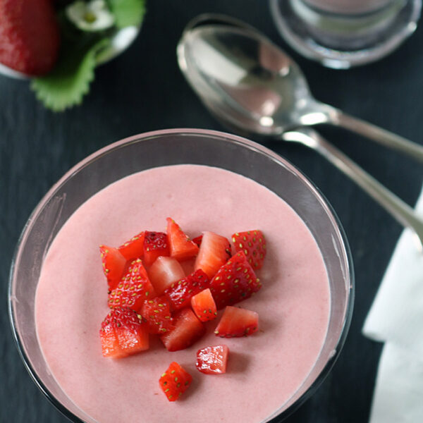 A glass dessert dish filled with pink strawberry mousse, topped with diced fresh strawberries. A spoon, napkin, and whole strawberries are placed nearby on a dark surface.