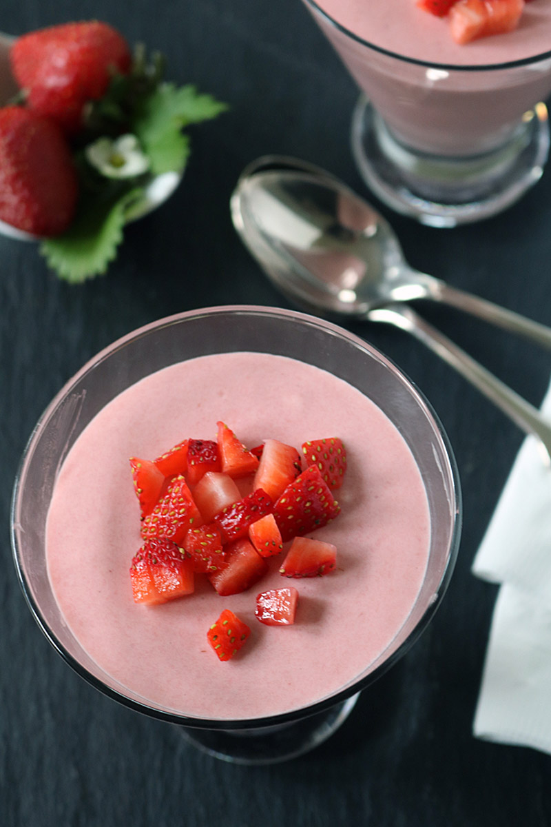 A glass dessert dish filled with pink strawberry mousse, topped with diced fresh strawberries. A spoon, napkin, and whole strawberries are placed nearby on a dark surface.