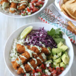 Two bowls filled with rice, crispy breaded fish, diced avocado, purple cabbage, pico de gallo, cilantro, and a lime wedge, topped with creamy sauce. Tortilla chips sit on the side with utensils on a napkin.