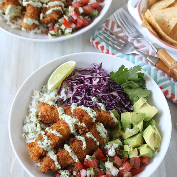 Two bowls filled with rice, crispy breaded fish, diced avocado, purple cabbage, pico de gallo, cilantro, and a lime wedge, topped with creamy sauce. Tortilla chips sit on the side with utensils on a napkin.