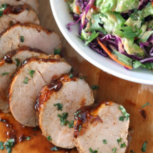 Sliced pork tenderloin with sauce on a wooden board, garnished with herbs, next to a bowl of colorful coleslaw made of shredded cabbage, carrots, and green vegetables.