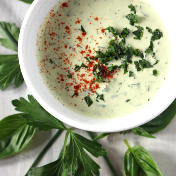 A bowl of creamy herb sauce garnished with chopped parsley and paprika sits on a white surface, surrounded by fresh parsley and basil leaves.