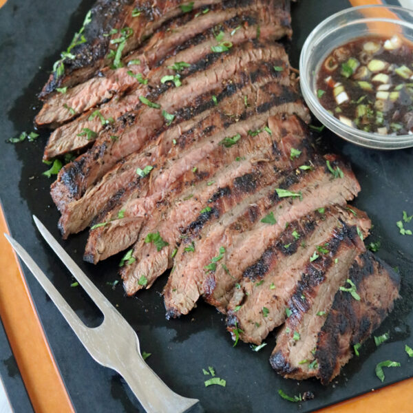 Sliced grilled steak garnished with chopped herbs on a black cutting board, served with a small bowl of dipping sauce containing green onions. A meat fork rests nearby.