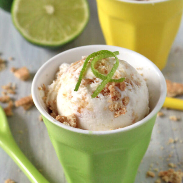 A scoop of ice cream sprinkled with crumbs and topped with lime zest sits in a green cup, with a green spoon nearby. A yellow cup with more ice cream and a halved lime are in the background.